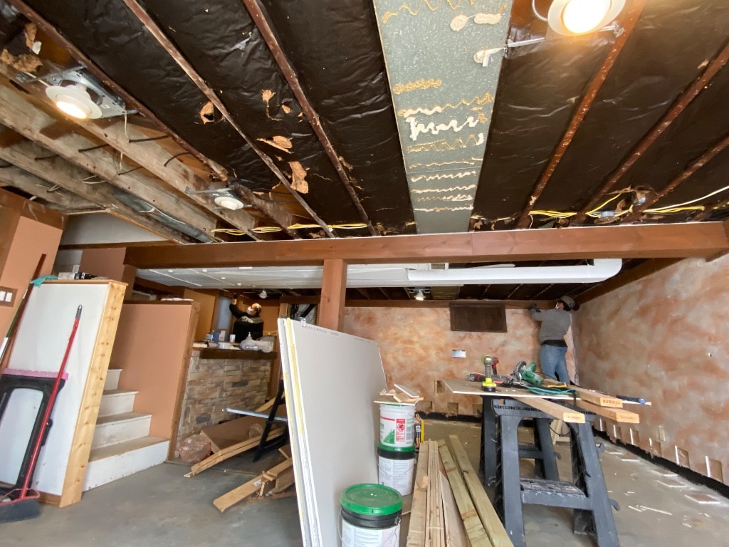 Basement gut renovation showing exposed joists, vapor barrier, drywall staged, and crew working on waterproofing and electrical rough-in