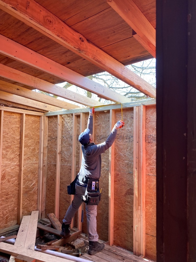 Set2 Go carpenter with tool belt reaching up with tape measure inside a wood-framed structure with OSB sheathing and exposed rafters