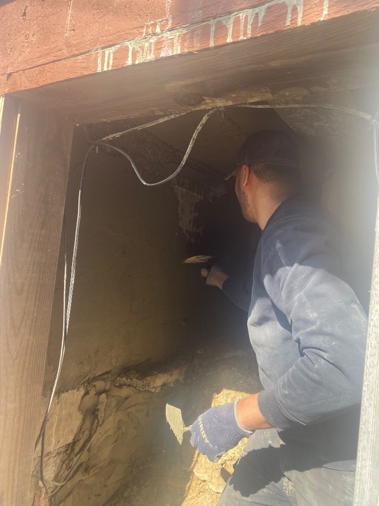Worker chiseling and troweling inside a foundation opening with exposed soil, block wall, and structural repair in progress