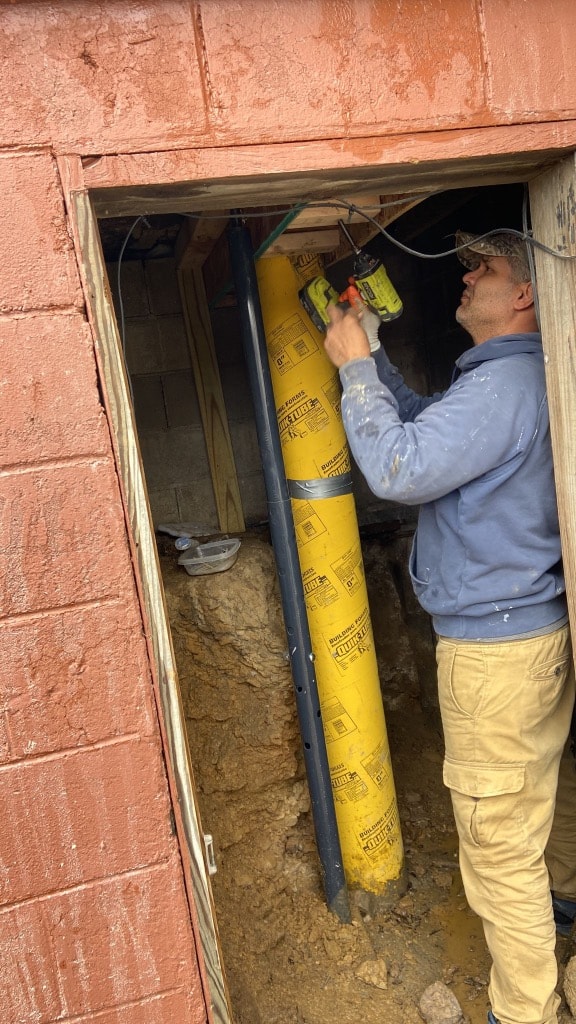 Worker drilling and setting a concrete Sonotube foundation pier inside a crawl space opening on a Pittsburgh brick home