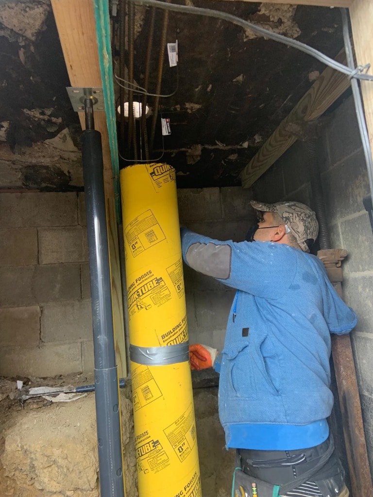 Worker positioning a concrete pier form inside a crawl space with exposed floor joists and block foundation wall visible
