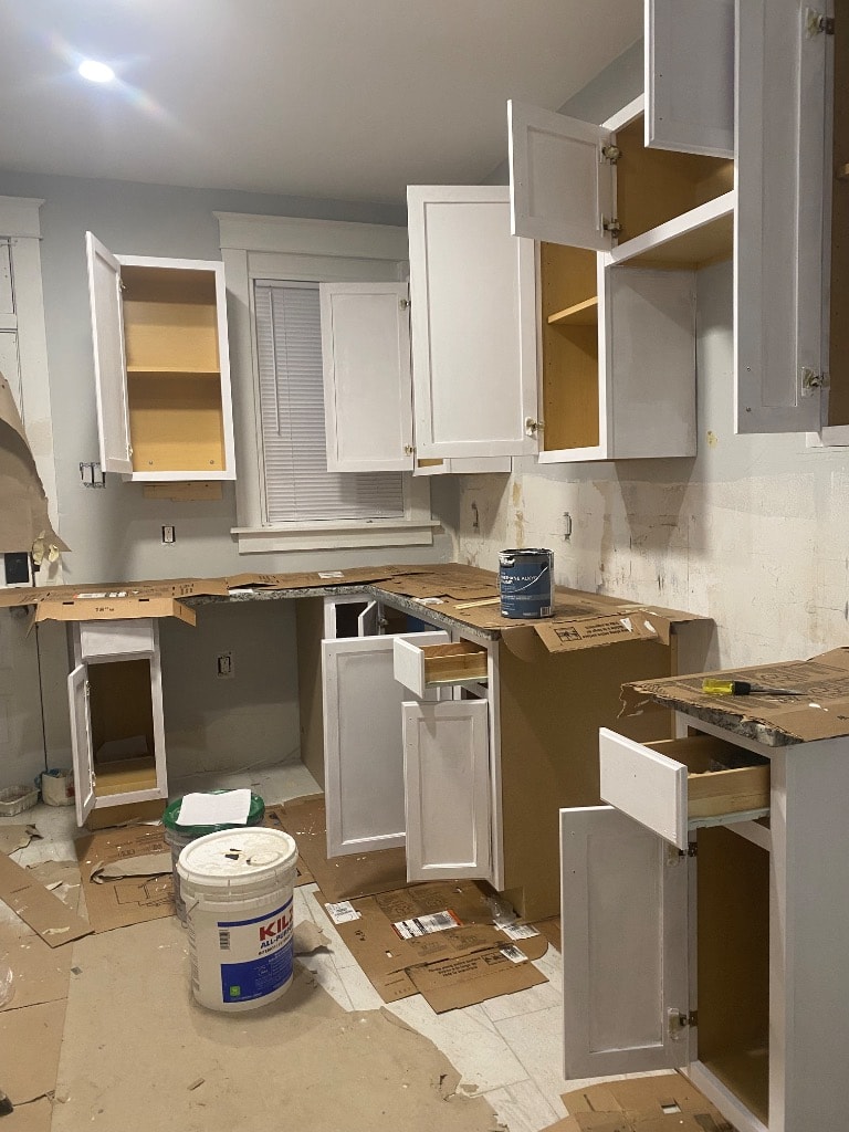 Kitchen renovation in progress with new white shaker cabinets being installed, countertops protected, and paint supplies on site
