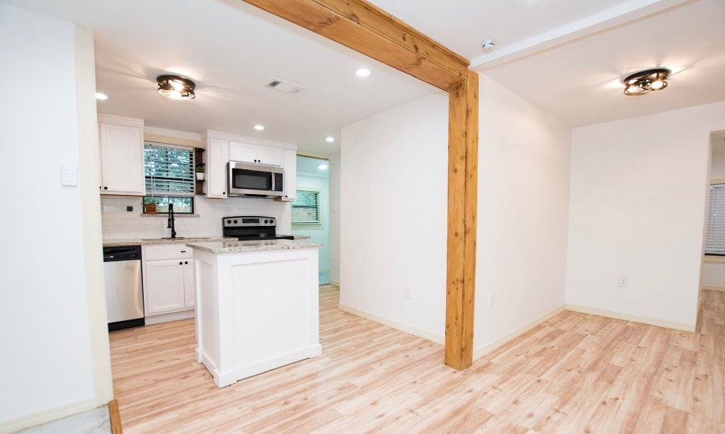 Completed kitchen remodel with white shaker cabinets, island, stainless appliances, light hardwood floors, and exposed wood beam accent